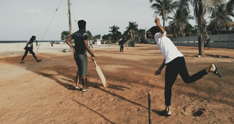 A group of young men playing cricket on a sandy beach under clear skies.